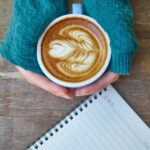 Person holding a coffee with latte art beside an open journal, preparing for autumn journaling for mental health and self-care.