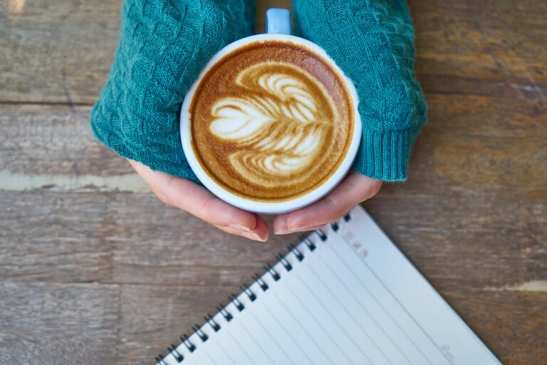 Person holding a coffee with latte art beside an open journal, preparing for autumn journaling for mental health and self-care.